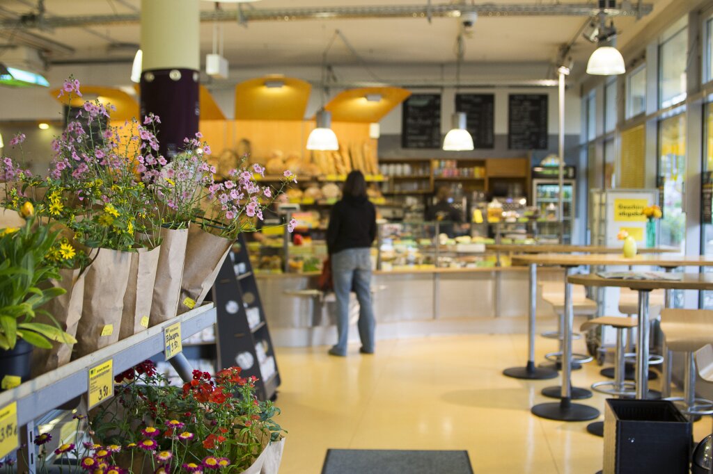 Blick in ein Café mit einer Theke im Hintergrund, an der eine Person steht; im Vordergrund links Blumen in Papierverpackung, rechts Stehtische und Stühle.