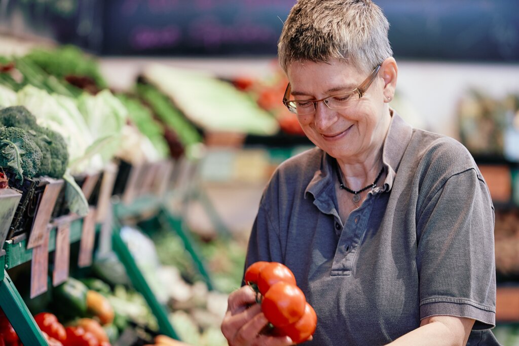 Eine Person mit kurzem grauen Haar und Brille hält lächelnd Tomaten in einem Gemüseladen.