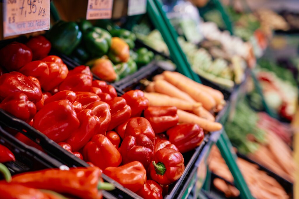 Verschiedene frische Gemüsesorten, darunter rote Paprika, grüne Paprika und Karotten, liegen in Auslagen eines Gemüsestands.