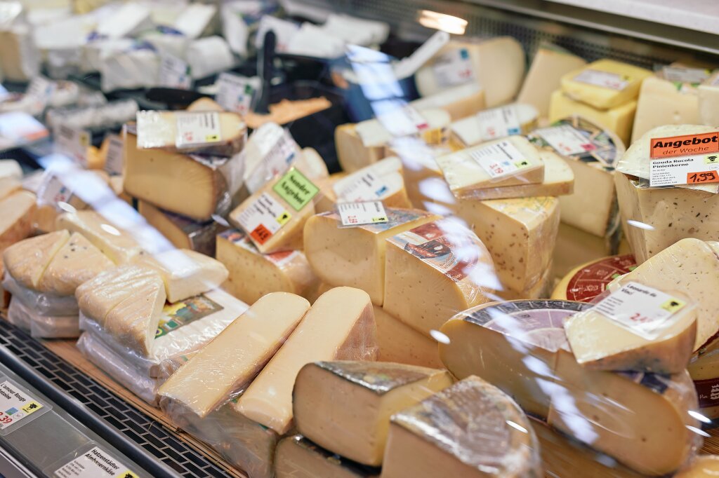 Verschiedene Sorten Käse liegen in einer Auslage hinter Glas, teilweise in Plastik verpackt und mit Preisschildern versehen.