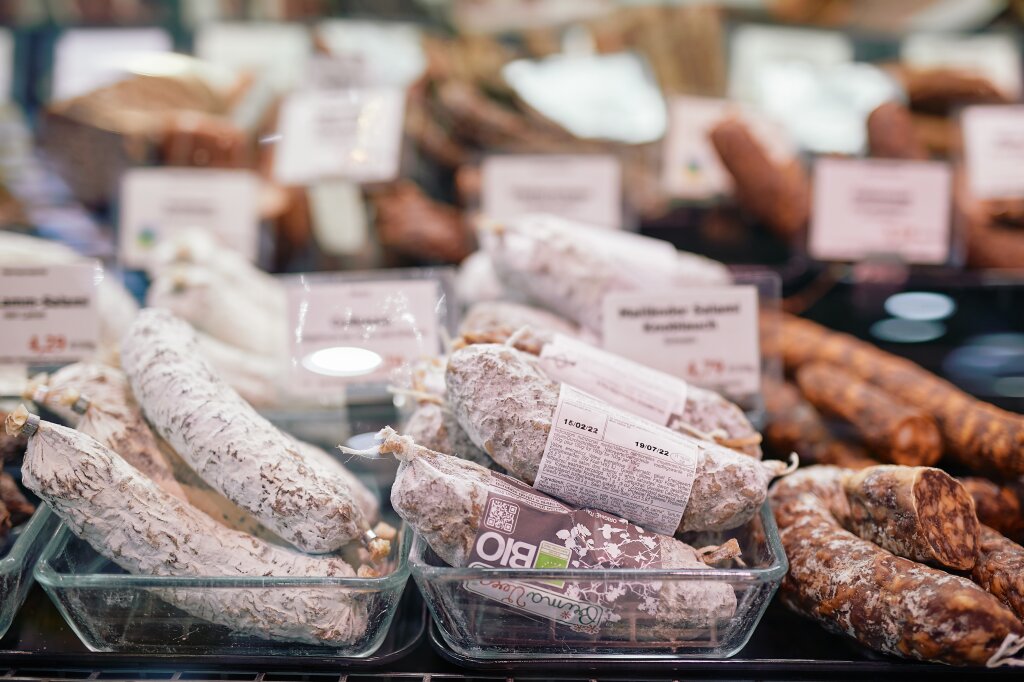 Verschiedene Wurstwaren liegen in Glasbehältern in einer Auslage, teils mit weißem Edelschimmel überzogen, dahinter befinden sich Preisschilder.