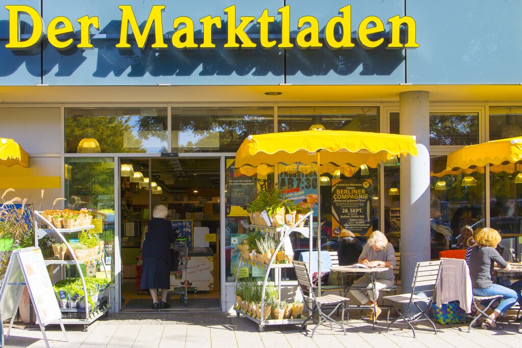 Eingang eines Ladens mit der Aufschrift „Der Marktladen“, gelbe Markisen, Blumenauslagen, Stühle und Tische draußen; eine ältere Frau betritt den Laden, zwei Frauen sitzen am Tisch.