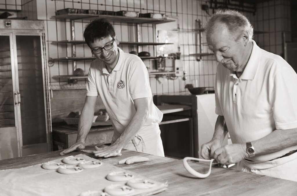 Zwei Männer in Bäckerkluft formen Brezeln an einem Holztisch in einer Backstube.