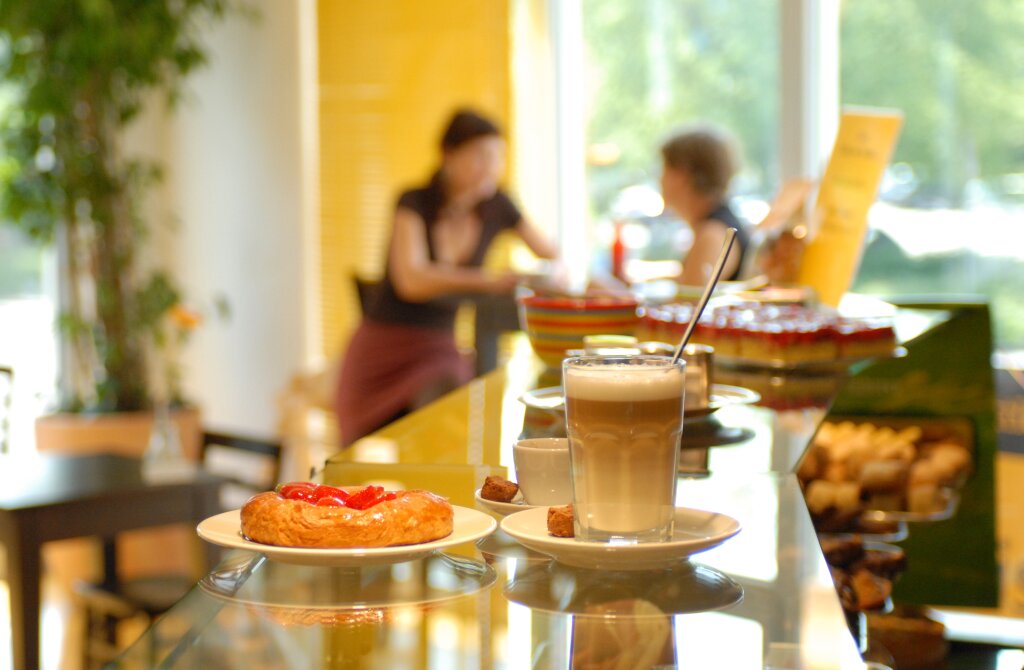 Ein Glas Latte Macchiato mit Löffel, ein Stück Gebäck mit Erdbeeren und ein kleiner Würfelzucker stehen auf einem Cafétresen, im Hintergrund sitzen unscharf zwei Personen an einem Tisch.