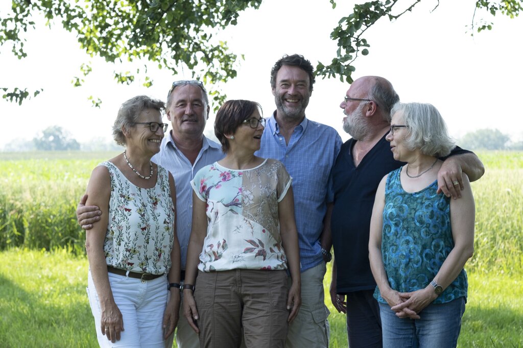 Sechs ältere Erwachsene stehen lachend im Freien auf einer Wiese, im Hintergrund grüne Bäume und Felder.